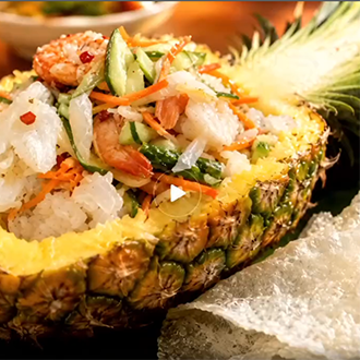 A vibrant Vietnamese seafood salad featuring shrimp, squid, green papaya, and mint leaves, beautifully served inside a hollowed-out pineapple boat next to pieces of crispy toasted rice paper.